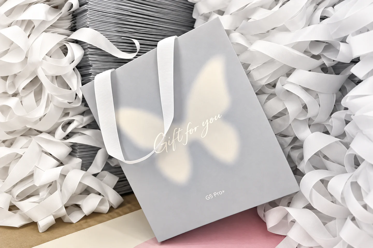 Physical Paper Bag Sample A grey paper bag with white ribbon handles resting on a stack of folded bags and loose ribbon.