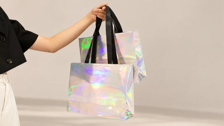 A person holding two unprinted holographic silver paper bags with rainbow reflections and broad black ribbon handles against a beige background