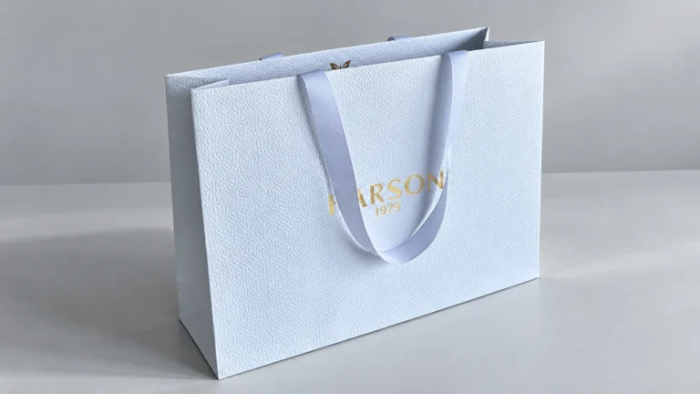 Pale blue textured paper bag with gold-coloured lettering, matching ribbon handles and a visible side gusset