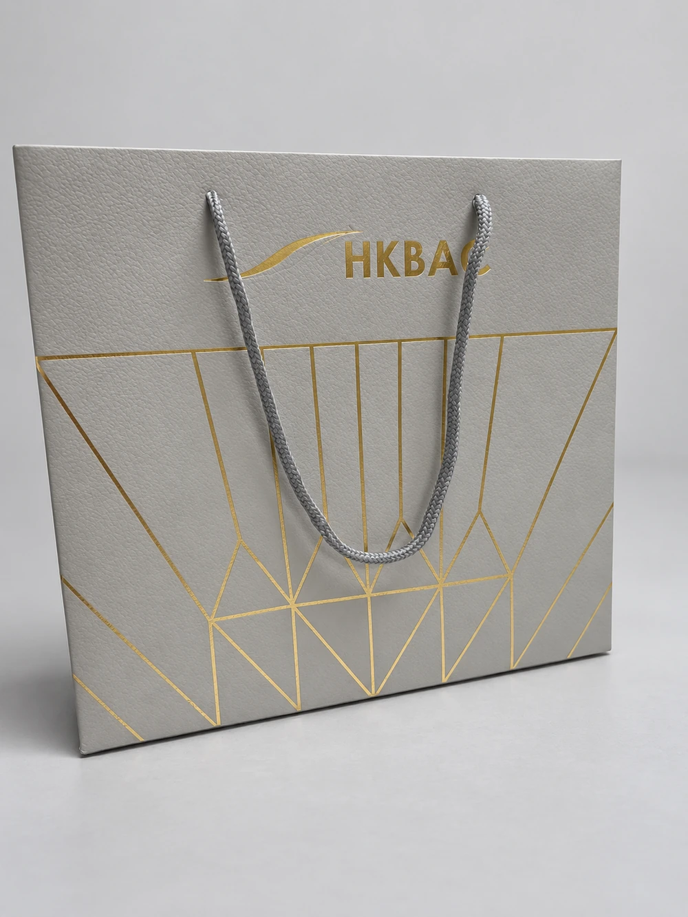 Grey textured paper bag with a grey braided cord handle, gold-coloured lettering and geometric lines