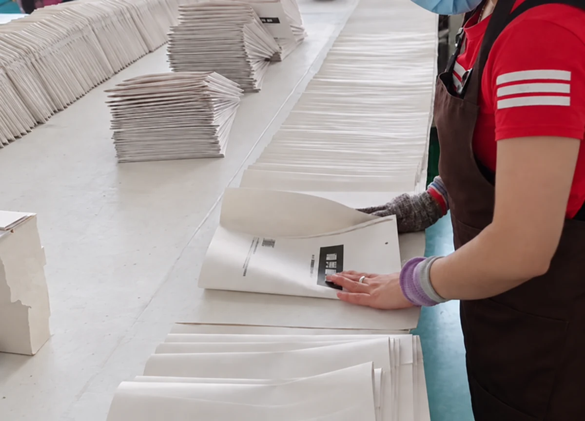 A worker handling white paper bags at a production table during manual finishing