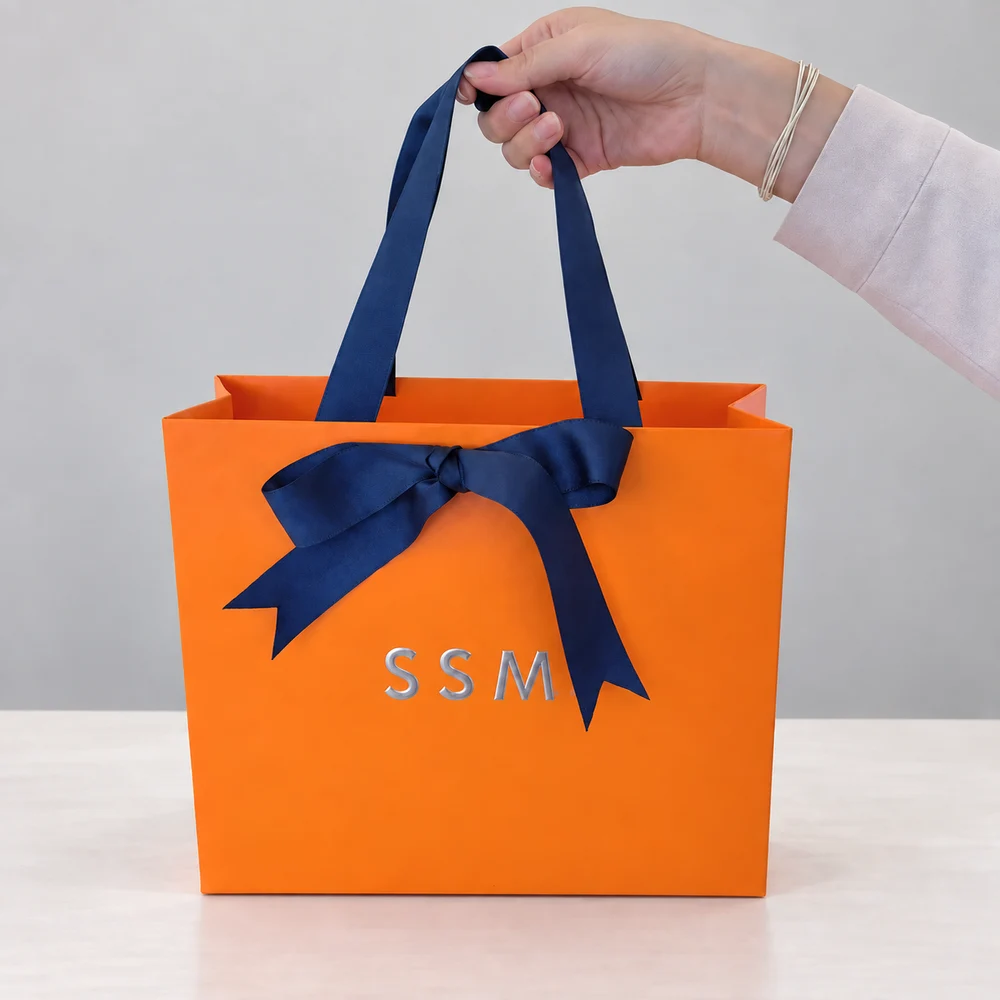 Orange SSM paper bag with blue ribbon handles and a bow, held above a light tabletop