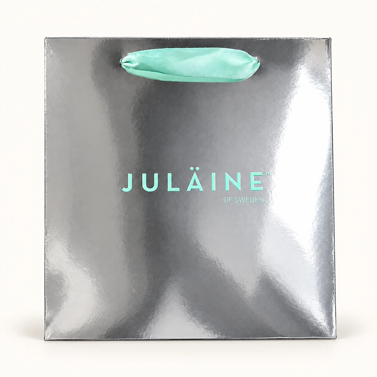 Reflective silver JULÄINE paper bag with mint-coloured lettering, OF SWEDEN text and a matching flat ribbon handle