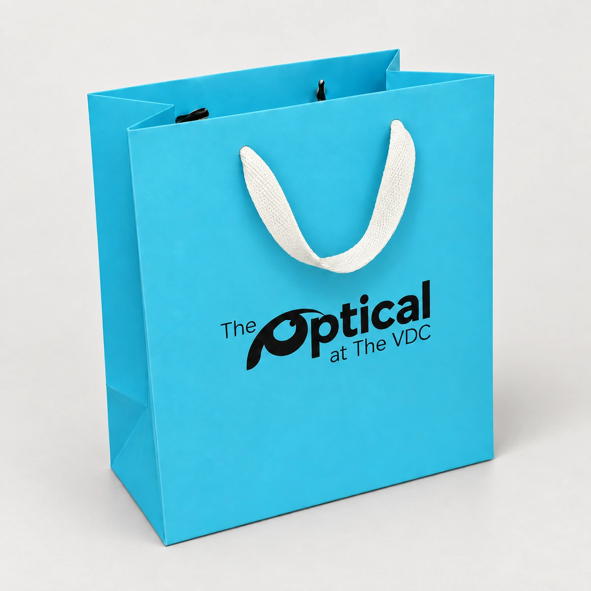 Turquoise The Optical paper bag with a hanging white ribbon handle and dark end fittings visible inside the top