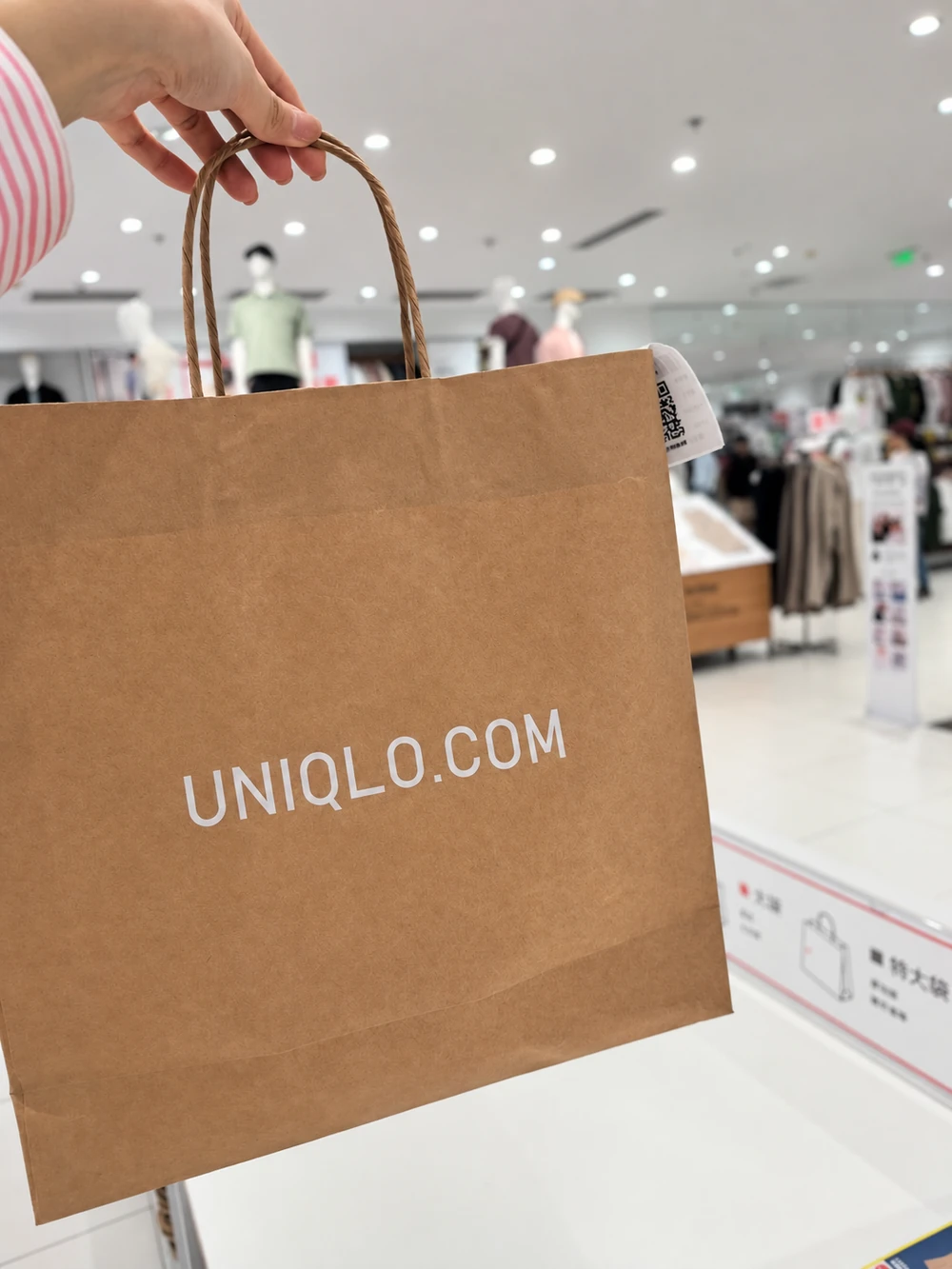 A hand holds the twisted handles of a brown shopping bag printed UNIQLO.COM inside a clothing store.