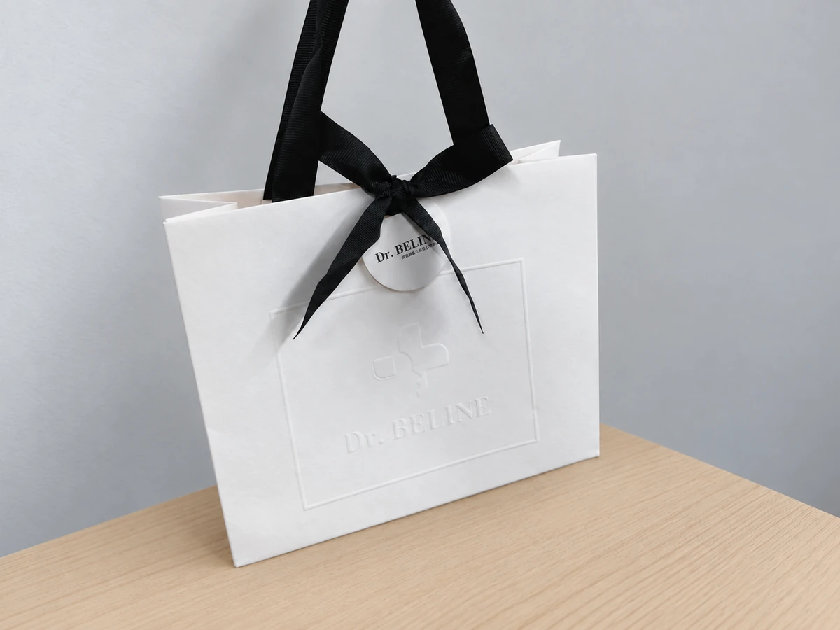 White Dr. BELINE paper bag with a subtle raised border and logo, black ribbon bow and round hanging tag