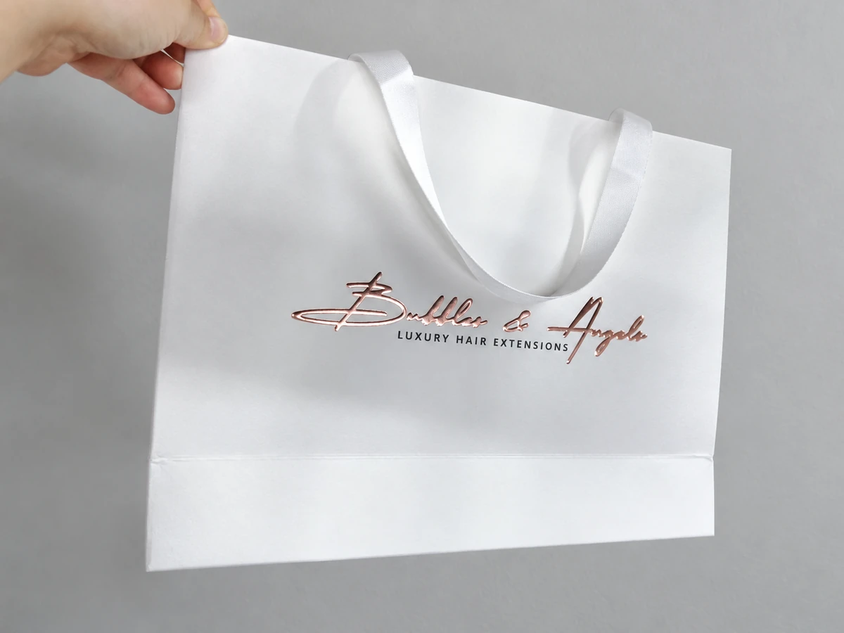 Hand-held white paper bag with a rose-gold-coloured script logo, black LUXURY HAIR EXTENSIONS lettering and a white ribbon handle
