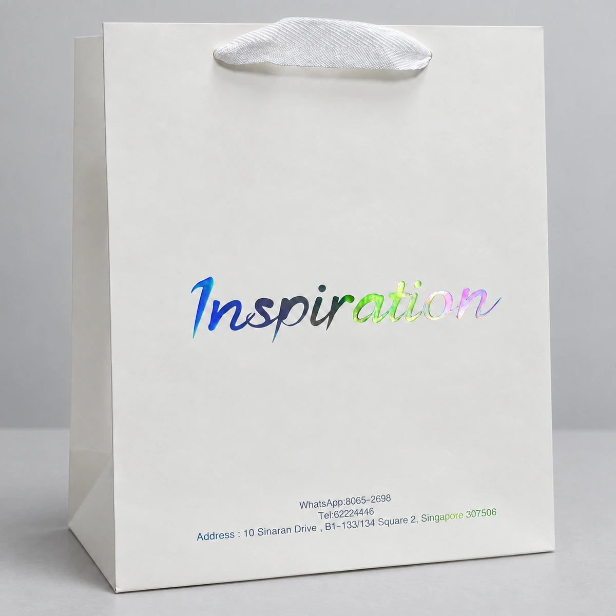 White paper bag with a ribbon handle and an Inspiration wordmark reflecting blue, green and pink