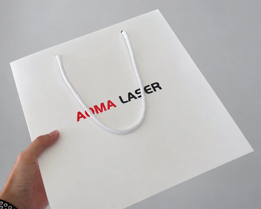 White paper bag with white rope handles and red AOMA and black LASER lettering