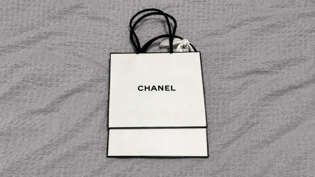 White CHANEL paper bag design with black borders, black rope handles and a small white ribbon displayed flat on grey textured fabric.