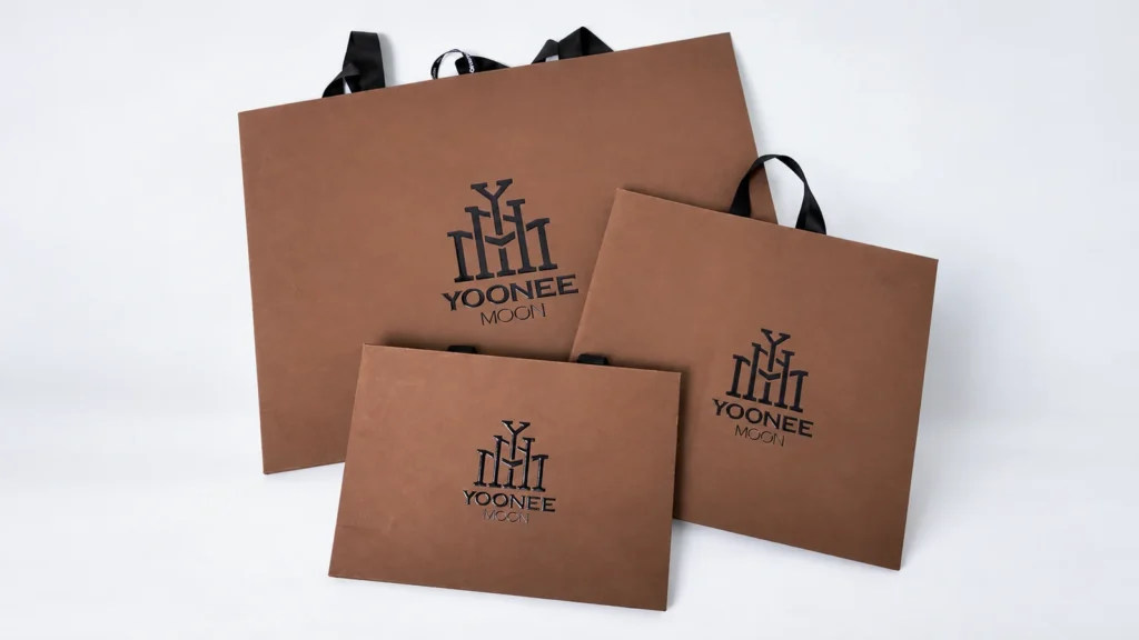 Three brown YOONEE MOON paper bags in different sizes with black ribbon handles.