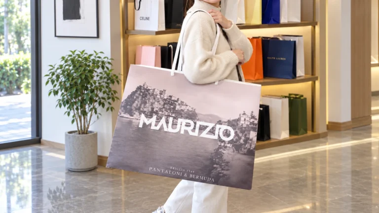 A person in a cream sweater carries a wide paper bag printed with a coastal landscape and a large white wordmark.