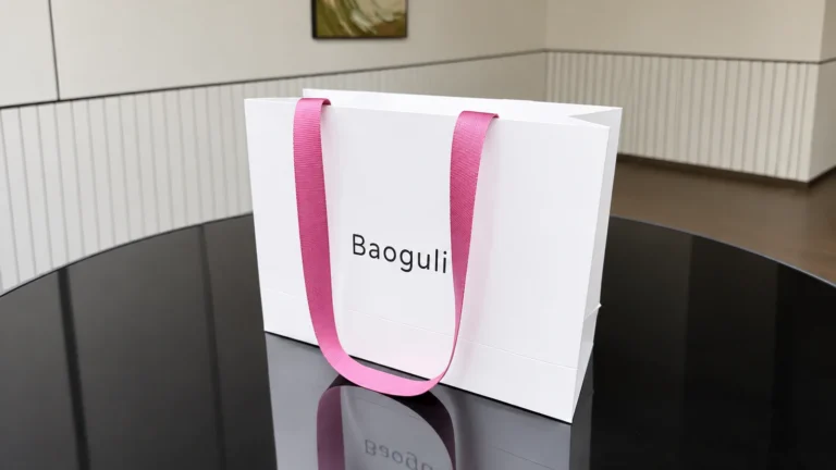 A white paper shopping bag with pink ribbed ribbon handles and a black wordmark stands on a dark reflective table.