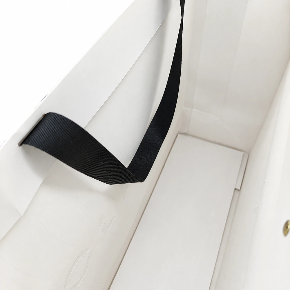 Inside a white paper bag, a flat white bottom card covers the base beneath a black ribbon handle.