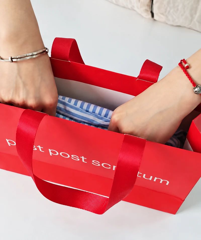 Hands placing a folded blue-and-white striped shirt inside a red paper shopping bag with ribbon handles.
