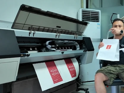 A digital printer produces a sheet with red artwork while a person beside it holds a white paper bag sample with a red logo.