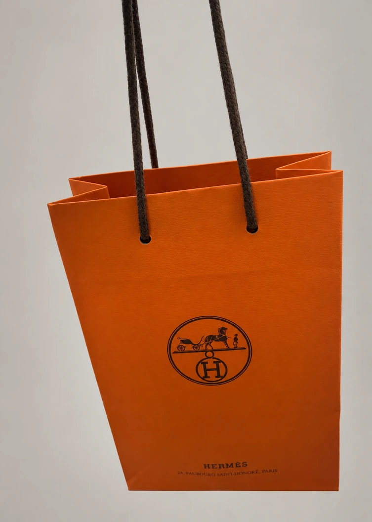 Tall orange Hermès paper bag with dark cord handles, a central emblem and small lettering near the base