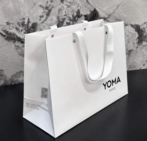 White YOMA SPACE paper bag with a clear front logo and blurred contact details and QR code on its side panel.