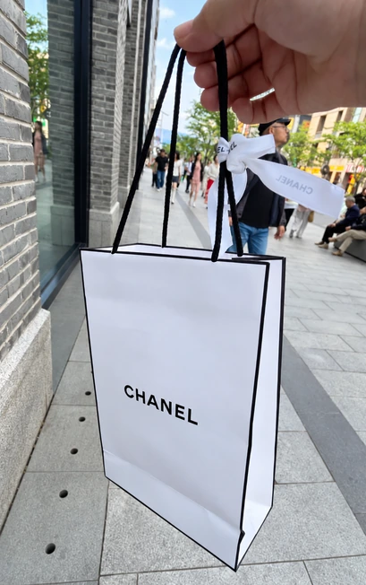 A hand holds a tall white CHANEL paper bag with a black border, long dark handles and a white ribbon on a street.