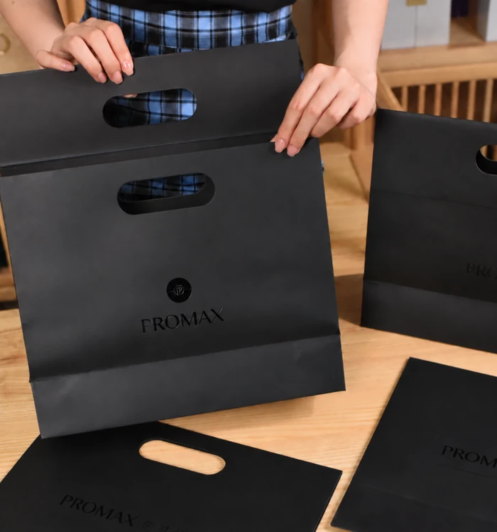 A person holds a black paper bag with an oval die-cut carrying opening above other black bags on a wooden table.