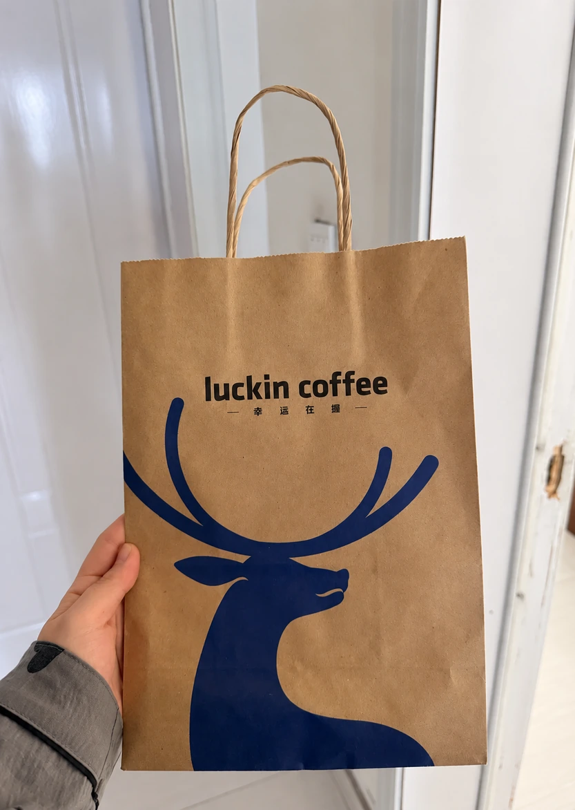 Brown Luckin Coffee paper bag with twisted handles, black lettering and a large blue deer graphic