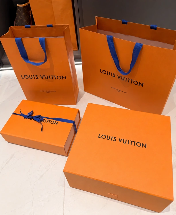 Two orange Louis Vuitton shopping bags with blue handles behind two matching orange gift boxes.