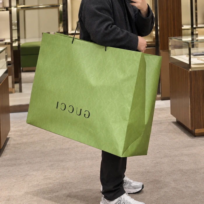 A person holds a broad green paper bag with a mottled surface, dark cord handles and a small dark wordmark.