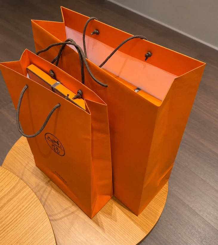 Two open orange Hermès paper bags showing dark cord ends tied into knots inside their folded tops