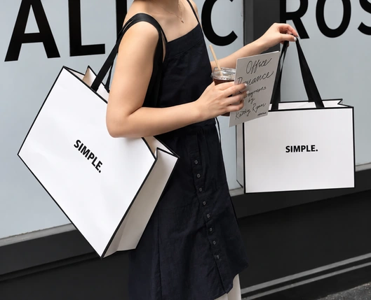 A person in a black dress carries two white SIMPLE. shopping bags with black borders and broad black handles.
