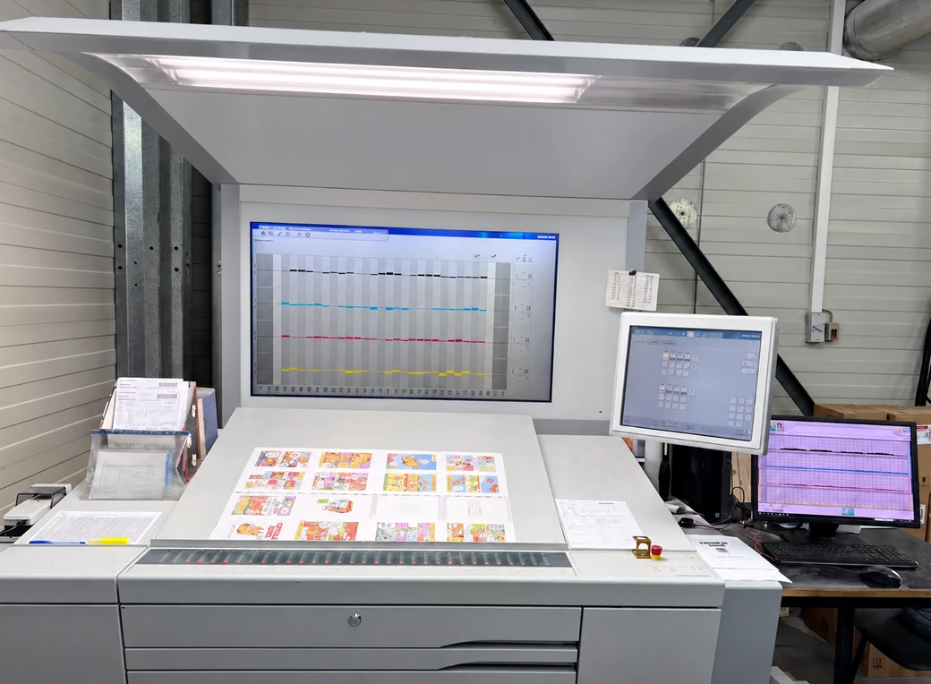 A multicolor printed sheet rests on an illuminated press control desk beside monitoring screens and inspection tools.