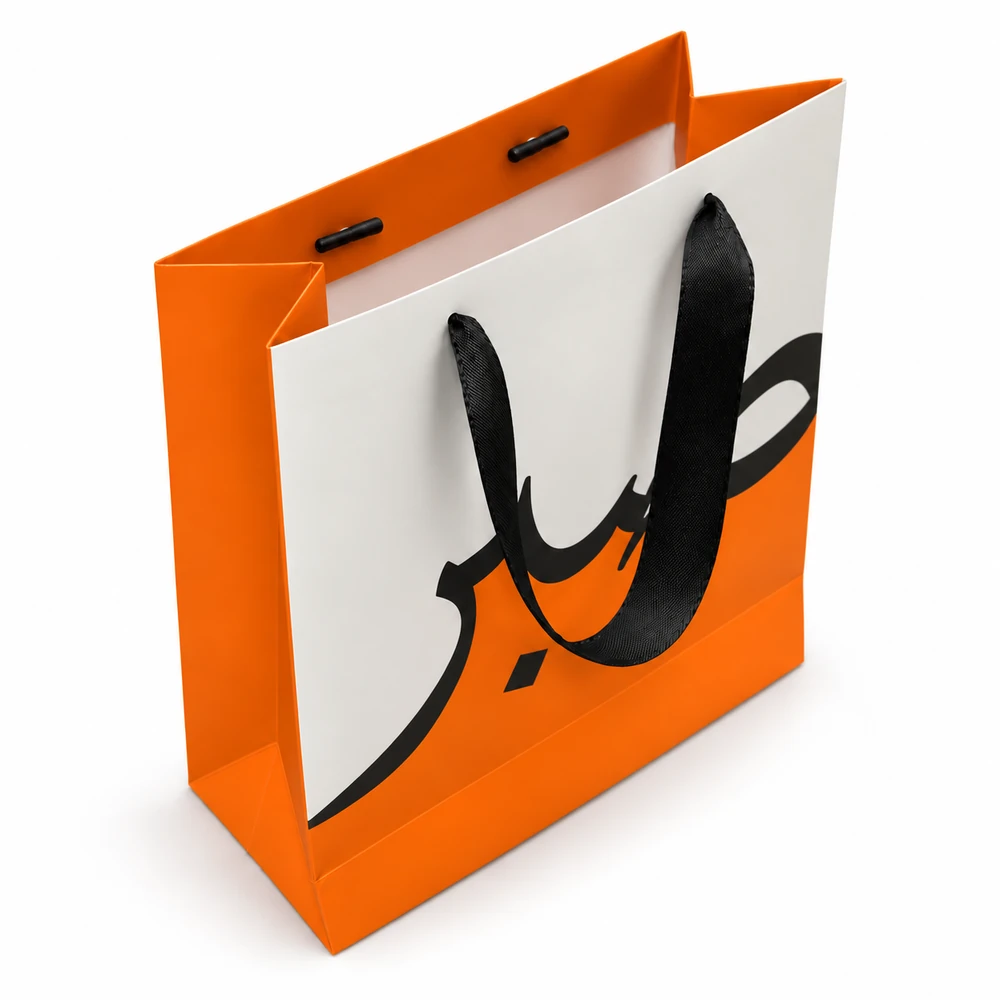 An orange-and-white paper bag has a black ribbon loop threaded through its front and two black handle end pieces visible inside the opposite top fold.