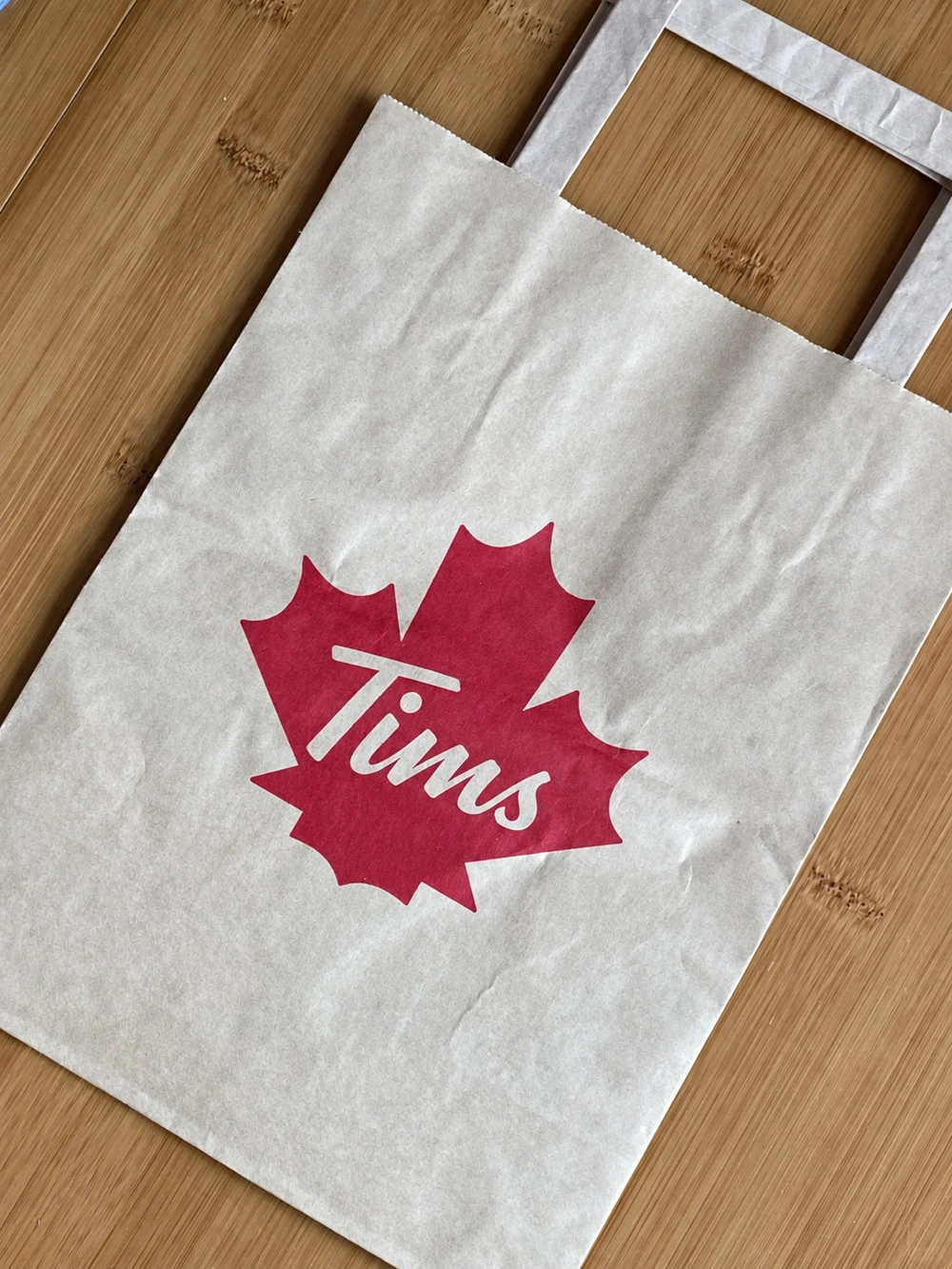 White paper bag with flat handles and a red maple-leaf graphic containing the white Tims wordmark
